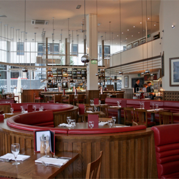 The interior at Piccolino. Bright, modern restaurant interior with large windows, high ceilings, and hanging pendant lights. The space features curved wooden booths with red leather seating and neatly set tables with glassware and menus. A long bar at the back is stocked with bottles, and the overall design combines warm wood tones with contemporary elements.