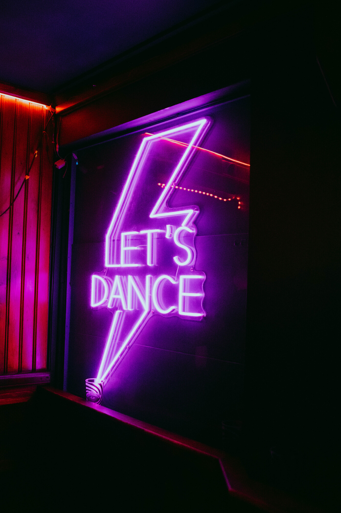 Pink and purple neon sign reading “Let’s Dance” inside a dimly lit bar or club space.