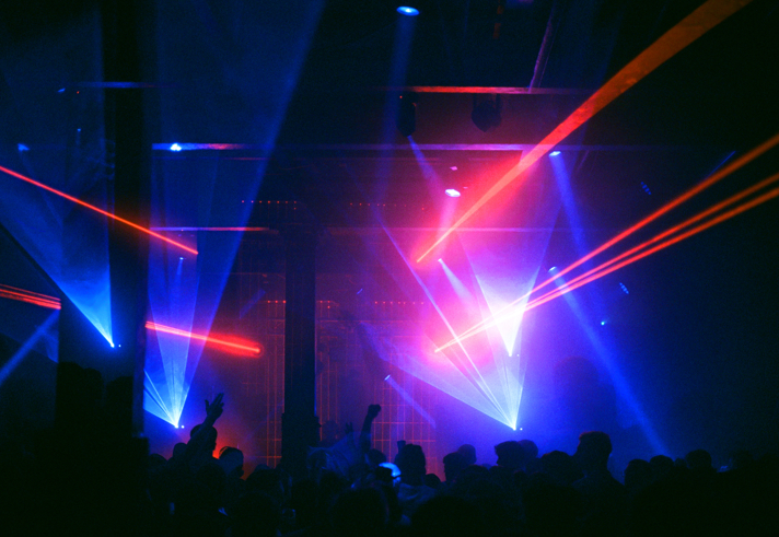 A crowded nightclub or music venue with vibrant laser lights in blue and red cutting across the dark space. Silhouettes of people are visible in the foreground, some with raised arms, creating a lively atmosphere. The beams of light converge toward the center stage area, highlighting the industrial-style ceiling and pillars. The overall scene conveys an energetic party or concert setting with dramatic lighting effects.