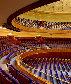 An elegant auditorium with tiered seating arranged in a semi-circular layout. The seats are upholstered in deep blue fabric, and the space features multiple balconies with matching seating. Warm lighting illuminates the curved wooden panels and the ornate, patterned ceiling. The overall design conveys a grand and formal atmosphere suitable for concerts, lectures, or large events.