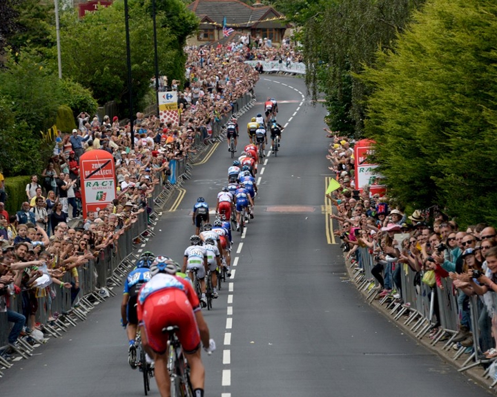 Downhill shot of the Tour De France coming through Sheffield, with crowds cheering them on either side of the road.