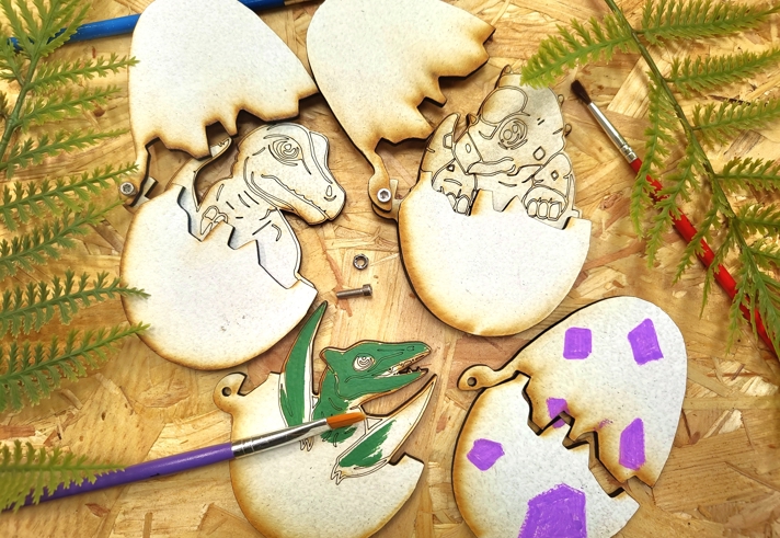 Three 2D wooden dinosaur eggs, that open to reveal a dinosaur, lie on a table ready to be decorated.