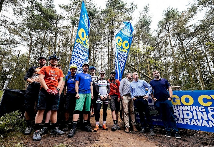 A group of people stands on a forest trail in front of two tall blue flags and a large banner that reads “GO CY” with words like “RUNNING,” “RIDING,” and “CARAVAN” visible. The individuals are dressed in casual and cycling gear, with some wearing helmets and gloves. The background shows tall trees and greenery, suggesting an outdoor cycling or adventure event.