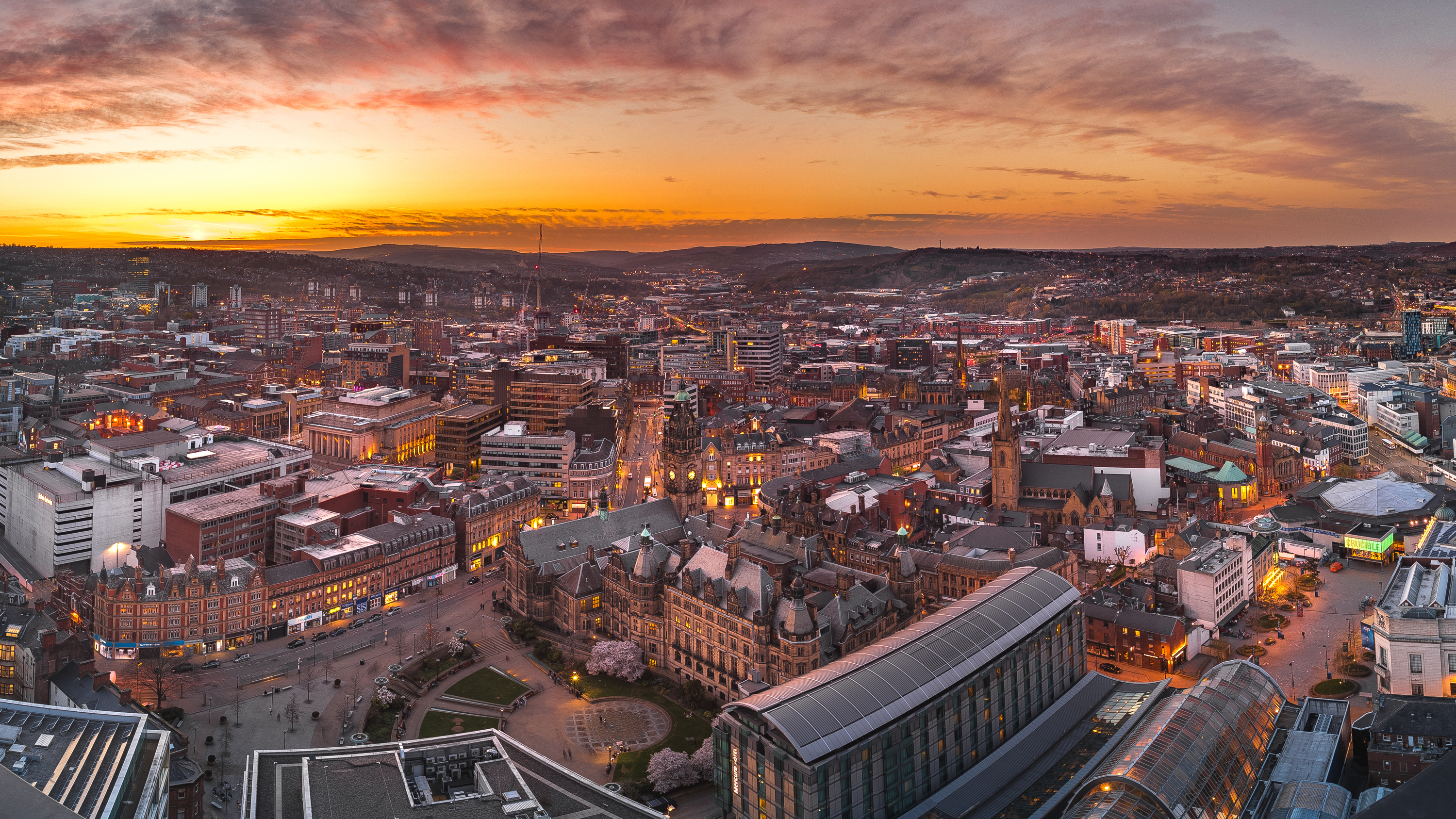 Panoramic view of Sheffield city centre at sunset, with warm orange and pink hues in the sky. Historic buildings and modern structures are visible, including the Town Hall and Winter Garden, surrounded by streets and rooftops stretching into the distant hills.
