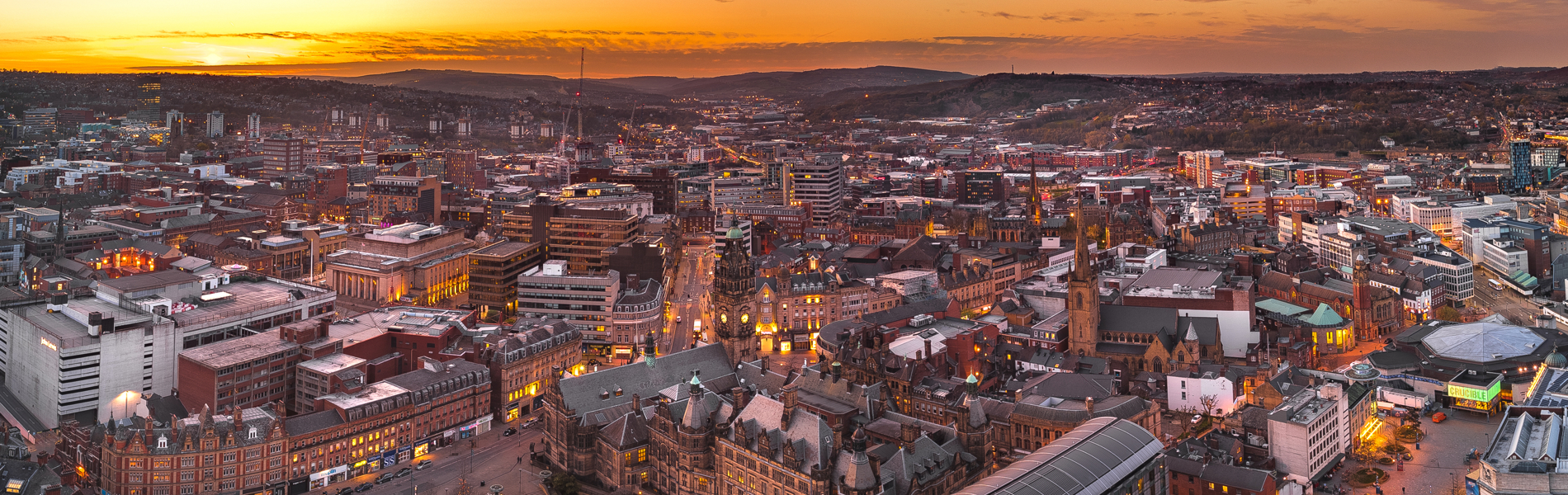 Panoramic view of Sheffield city centre at sunset, with warm orange and pink hues in the sky. Historic buildings and modern structures are visible, including the Town Hall and Winter Garden, surrounded by streets and rooftops stretching into the distant hills.