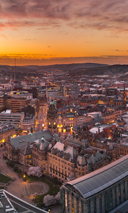 Panoramic view of Sheffield city centre at sunset, with warm orange and pink hues in the sky. Historic buildings and modern structures are visible, including the Town Hall and Winter Garden, surrounded by streets and rooftops stretching into the distant hills.