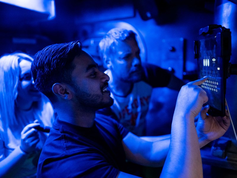 Three people in a room dimly lit by blue light are solving a puzzle at The Great Escape.