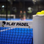 Close-up view of a padel court net with a white banner that reads “PLAY PADEL” in bold black letters and a logo on the left side. The net is black and attached to a padded post on the right, which has several yellow tennis balls resting on top. The court surface is bright blue, and the background is blurred, showing two players and parts of the indoor facility.