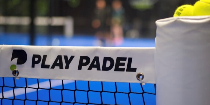 Close-up view of a padel court net with a white banner that reads “PLAY PADEL” in bold black letters and a logo on the left side. The net is black and attached to a padded post on the right, which has several yellow tennis balls resting on top. The court surface is bright blue, and the background is blurred, showing two players and parts of the indoor facility.