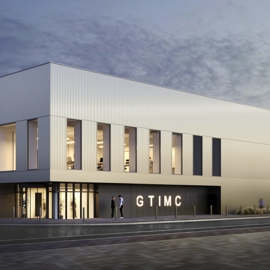 The exterior of the Gene Therapy Centre building. Modern two-story building with a sleek, minimalist design featuring vertical metal panels and large rectangular windows. The structure is illuminated from inside, creating a warm glow against the twilight sky. The entrance has glass doors and signage reading “GTIMC.” A few people are visible near the entrance, and blurred motion suggests activity in the foreground.