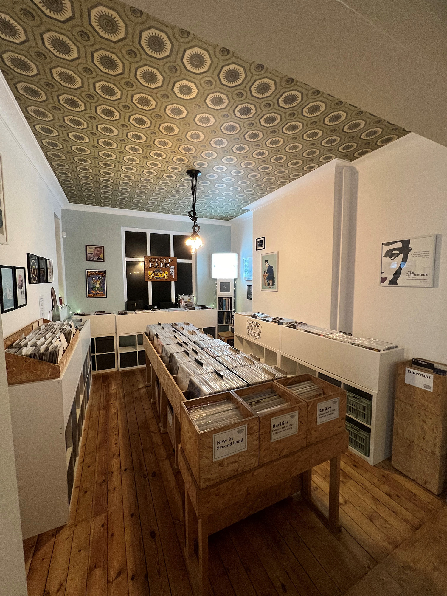 A cozy record shop interior with wooden floors and patterned ceiling tiles. Wooden crates and white shelving units hold vinyl records arranged neatly. Framed artwork and posters decorate the walls, and a single hanging light fixture illuminates the space.