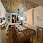 A cozy record shop interior with wooden floors and patterned ceiling tiles. Wooden crates and white shelving units hold vinyl records arranged neatly. Framed artwork and posters decorate the walls, and a single hanging light fixture illuminates the space.