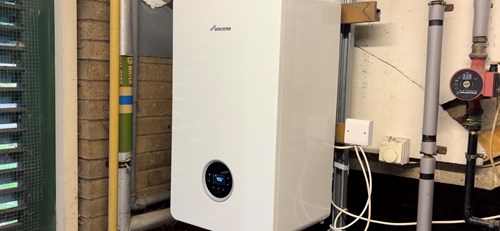 A white wall-mounted boiler unit with a digital display installed in a utility area. The boiler is connected to visible pipes and wiring, including a yellow gas pipe and a red valve on the right. The surrounding walls are brick and painted surfaces, and there is a metal conduit running vertically beside the boiler.