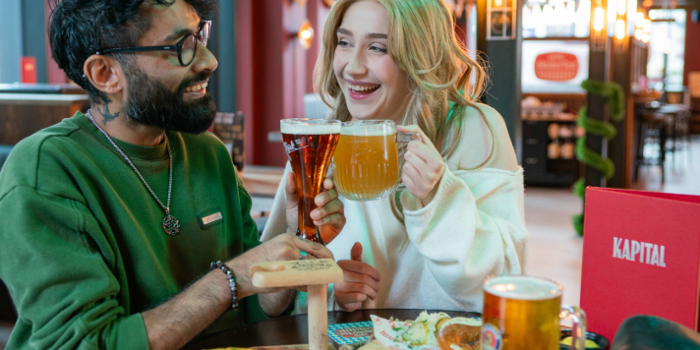 Two people are sitting at a table in a beer hall, clinking glasses of beer in a toast. One glass is a tall, dark beer and the other is a large mug of pale beer. The table has wooden boards with soft pretzels, mustard dip, and branded paper. A red menu with “KAPITAL” is visible, and the background features colourful bunting, warm lighting, and a modern interior with booths and bar seating.