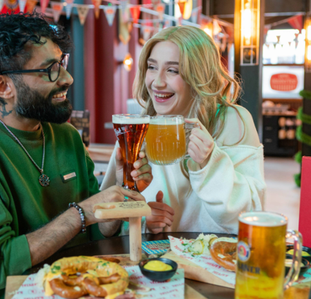 Two people are sitting at a table in a beer hall, clinking glasses of beer in a toast. One glass is a tall, dark beer and the other is a large mug of pale beer. The table has wooden boards with soft pretzels, mustard dip, and branded paper. A red menu with “KAPITAL” is visible, and the background features colourful bunting, warm lighting, and a modern interior with booths and bar seating.