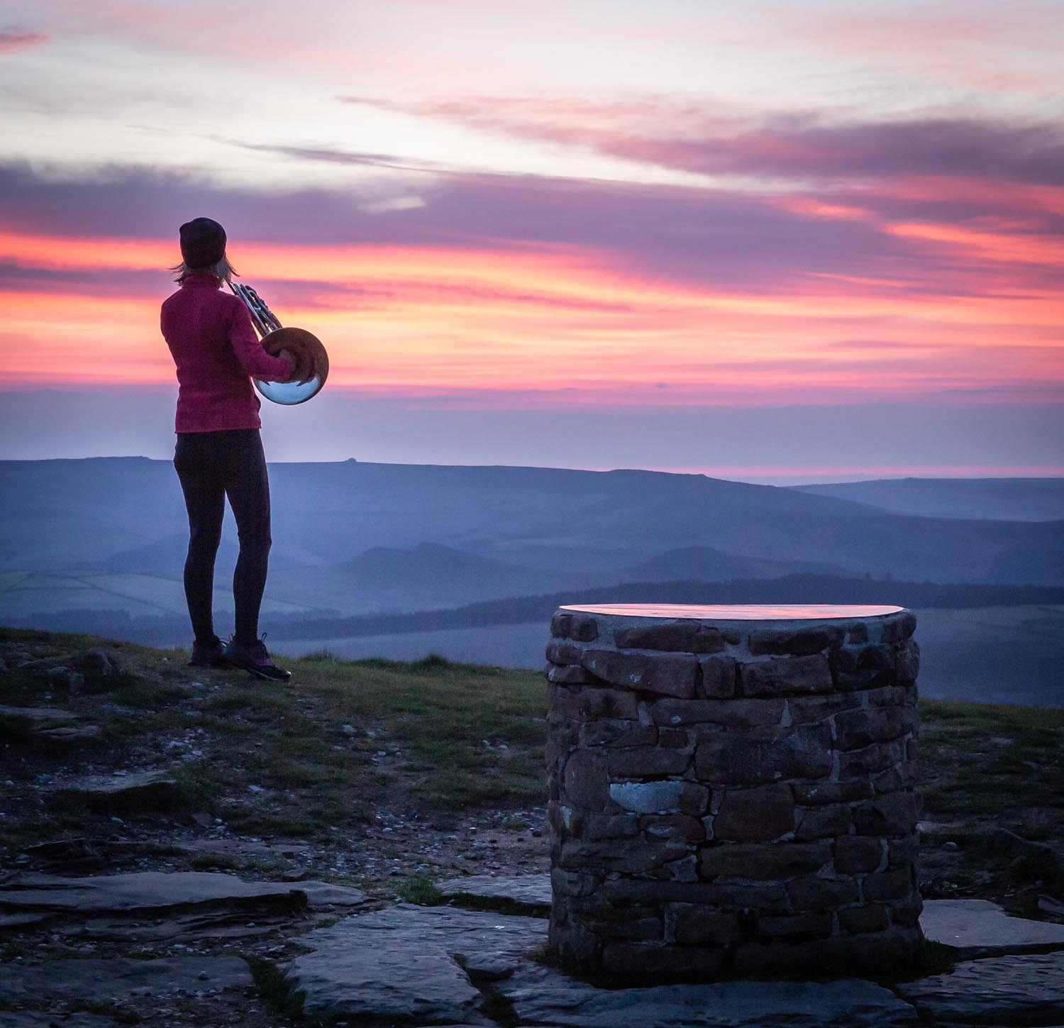 A person standing on a rocky hilltop at sunrise, holding a brass instrument while looking toward the horizon. The sky is filled with vibrant shades of pink, orange, and purple, casting a soft glow over the rolling hills in the distance. In the foreground, a circular stone structure sits on the ground, adding texture to the natural landscape.