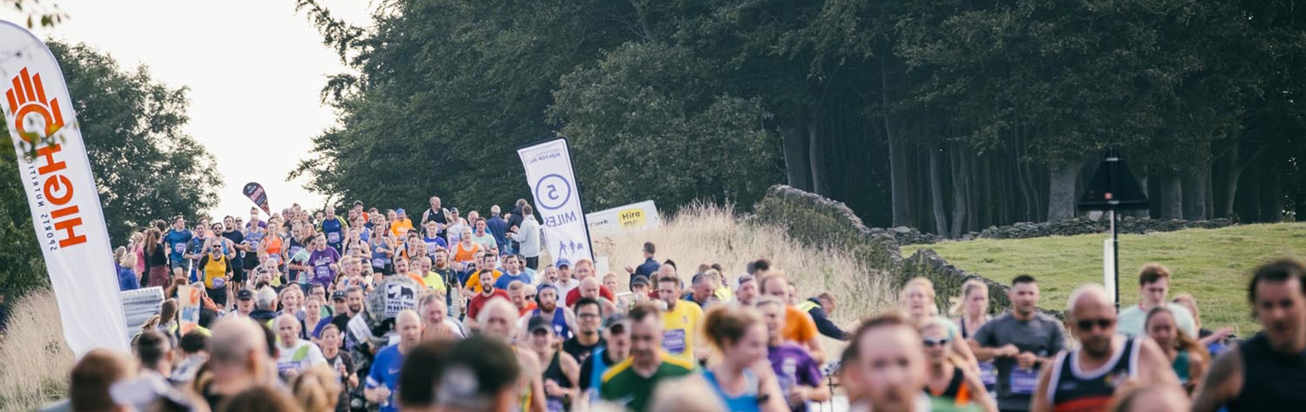 A large crowd is watching and cheering on runners running on a road. In the background is a large wood.