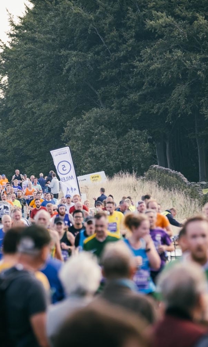 A large crowd is watching and cheering on runners running on a road. In the background is a large wood.