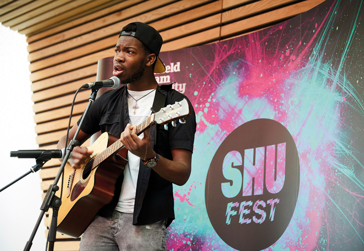 A person is performing on stage, playing an acoustic guitar and singing into a microphone. The backdrop features a colorful design with the text “SHU FEST” and “Sheffield Hallam University.” The setting appears to be an indoor event space with wooden panelling and stage lighting.