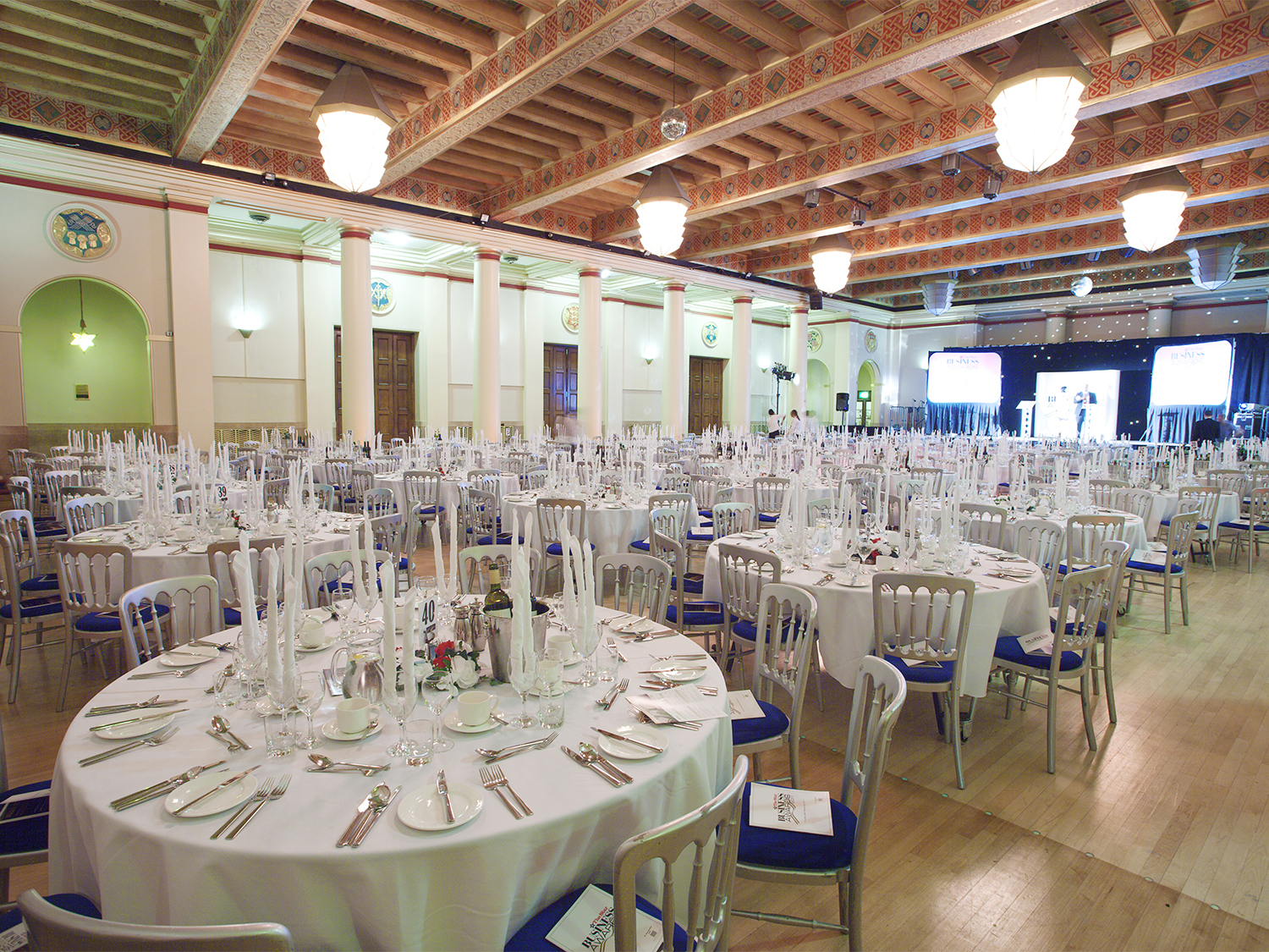 Elegant banquet hall set up for a formal event, featuring round tables covered with white tablecloths, neatly arranged silverware, glassware, and tall white candles. The room has high ceilings with exposed wooden beams, ornate light fixtures, and decorative columns, with a stage and large screens visible at the far end.