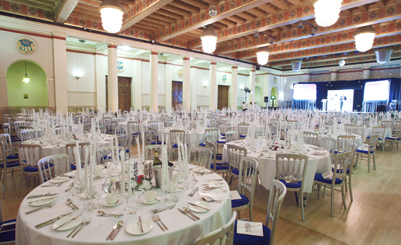 Elegant banquet hall set up for a formal event, featuring round tables covered with white tablecloths, neatly arranged silverware, glassware, and tall white candles. The room has high ceilings with exposed wooden beams, ornate light fixtures, and decorative columns, with a stage and large screens visible at the far end.