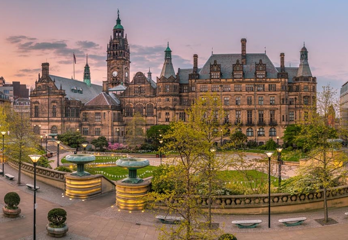 The Peace Gardens in Sheffield city centre; Panoramic view of a historic city square at sunset, featuring a large ornate building with towers and detailed architecture, likely a town hall. The square includes landscaped areas with trees, benches, and tiered fountains. Modern buildings border the scene, and the sky glows with warm orange and pink tones.