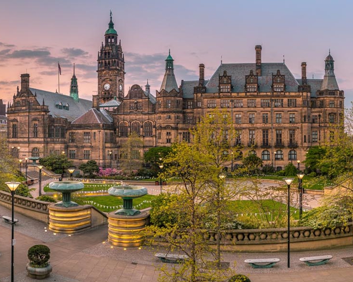 The Peace Gardens in Sheffield city centre; Panoramic view of a historic city square at sunset, featuring a large ornate building with towers and detailed architecture, likely a town hall. The square includes landscaped areas with trees, benches, and tiered fountains. Modern buildings border the scene, and the sky glows with warm orange and pink tones.