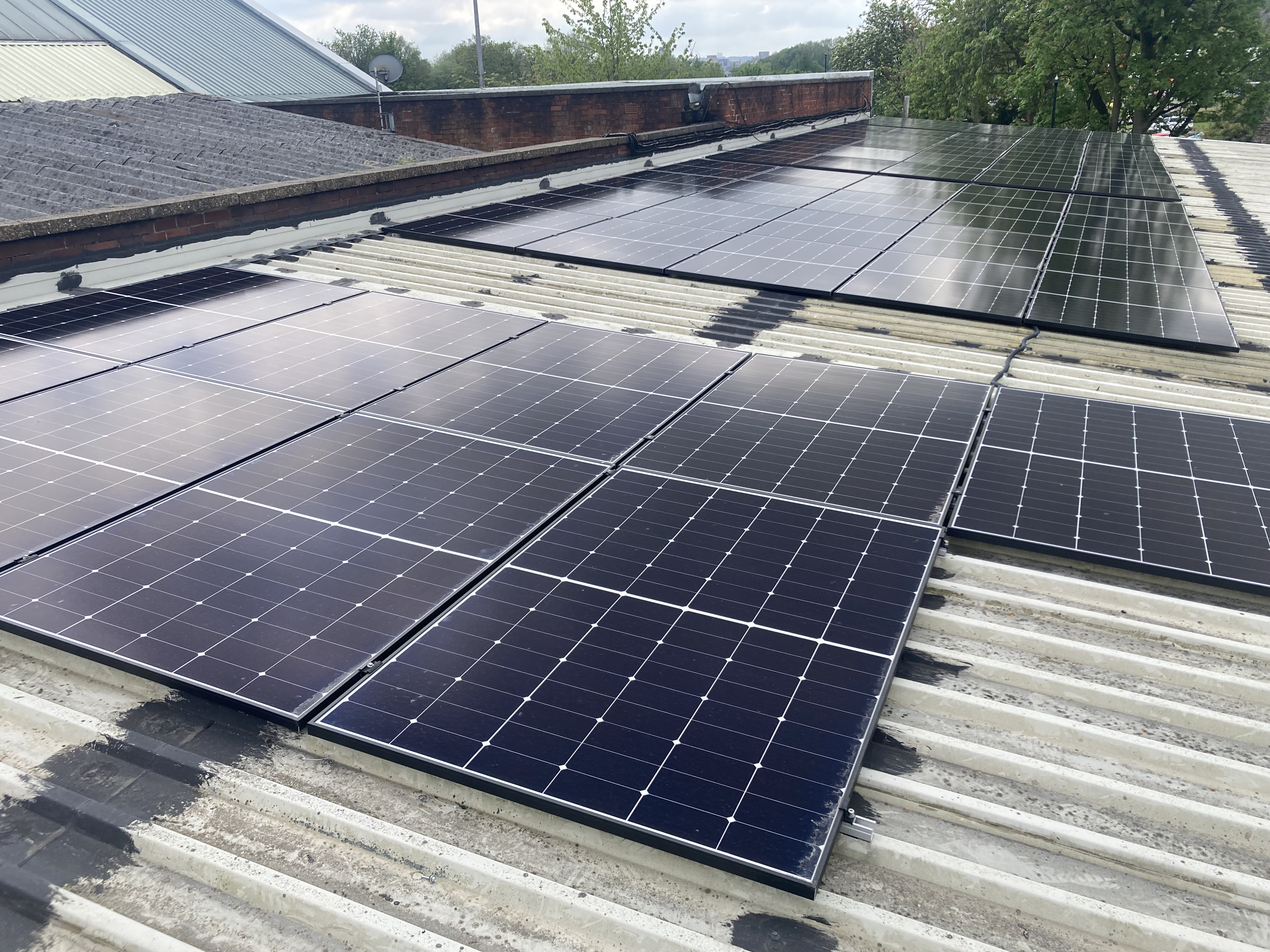 Rows of black solar panels installed on a corrugated metal rooftop. The panels are arranged in neat grids, reflecting light under a cloudy sky. In the background, there are additional rooftops, trees, and a glimpse of the horizon.