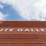 Exterior view of SITE Gallery’s brick building with its name displayed on the wall against a bright blue sky.