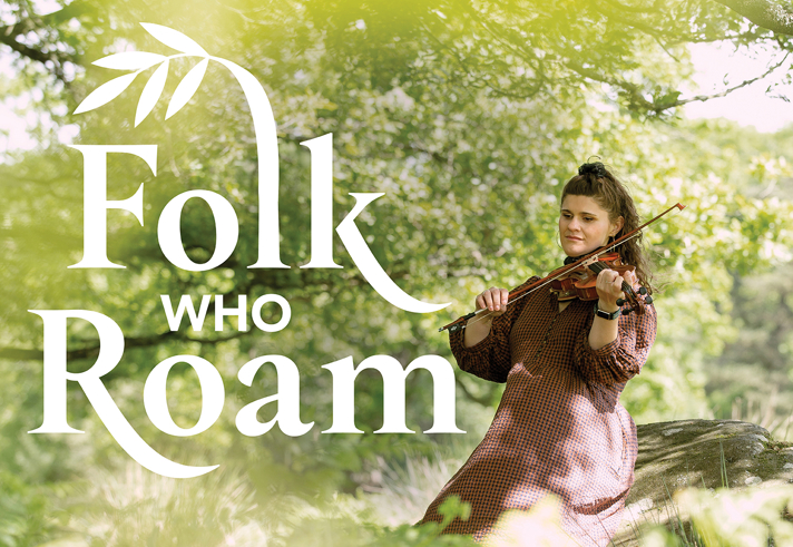A person wearing a long, patterned dress sits outdoors on a fallen tree trunk, playing a violin in a lush, green woodland setting. Sunlight filters through the leaves, creating a soft, natural glow. Large stylized text reading “Folk Who Roam” appears on the left side of the image, with a decorative leaf motif integrated into the lettering. Credit: Cat Vinton