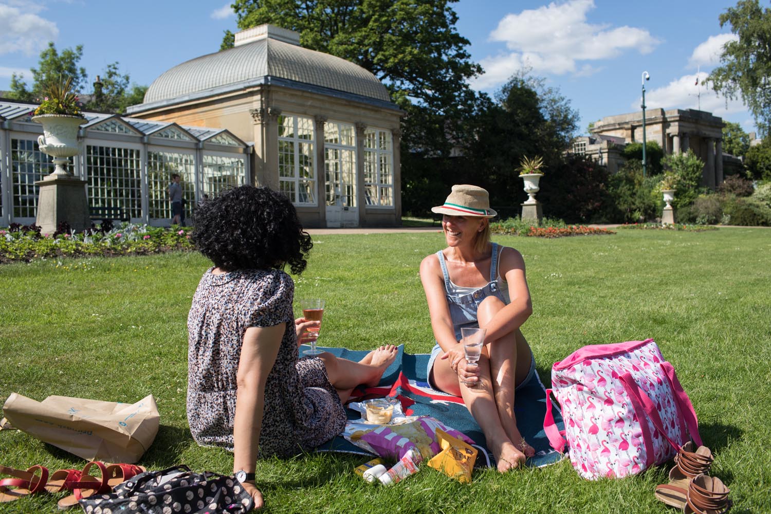 Two women are sat on the grass, on a bright, sunny day, at The Sheffield Botanical Gardens having a picnic and chatting. 