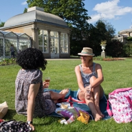 Two women are sat on the grass, on a bright, sunny day, at The Sheffield Botanical Gardens having a picnic and chatting. 