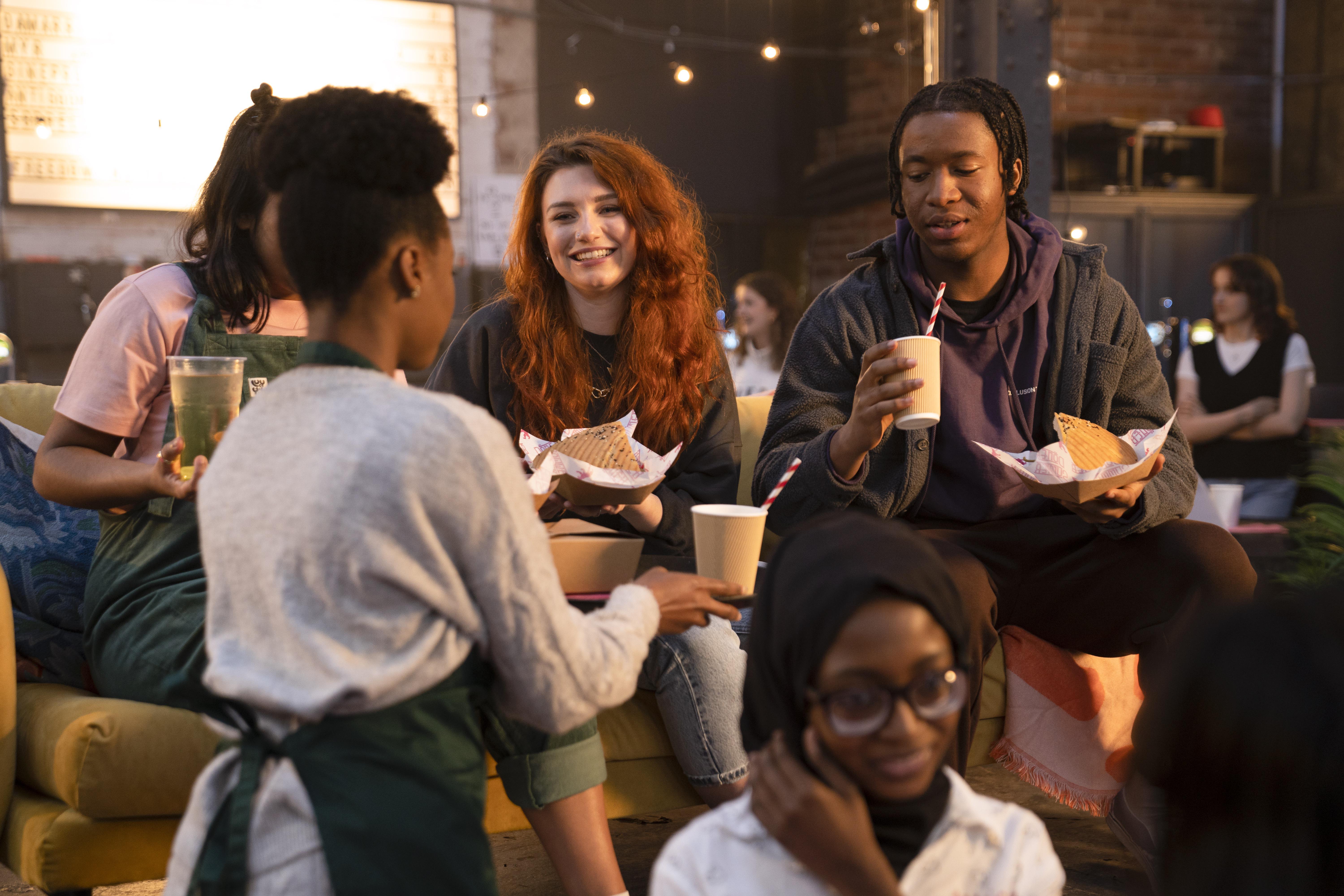 A cozy indoor setting with several people gathered on a sofa and nearby chairs, holding paper cups and food trays. Warm string lights hang overhead, and the background shows exposed brick walls and soft ambient lighting, creating a relaxed social atmosphere.