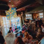  A crowded live music performance inside the Frog & Parrot pub, with warm lighting, patterned ceilings and a band playing near the front windows while people gather closely to watch.