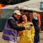 Two people standing near a festival stall, one holding a phone and the other a snack.