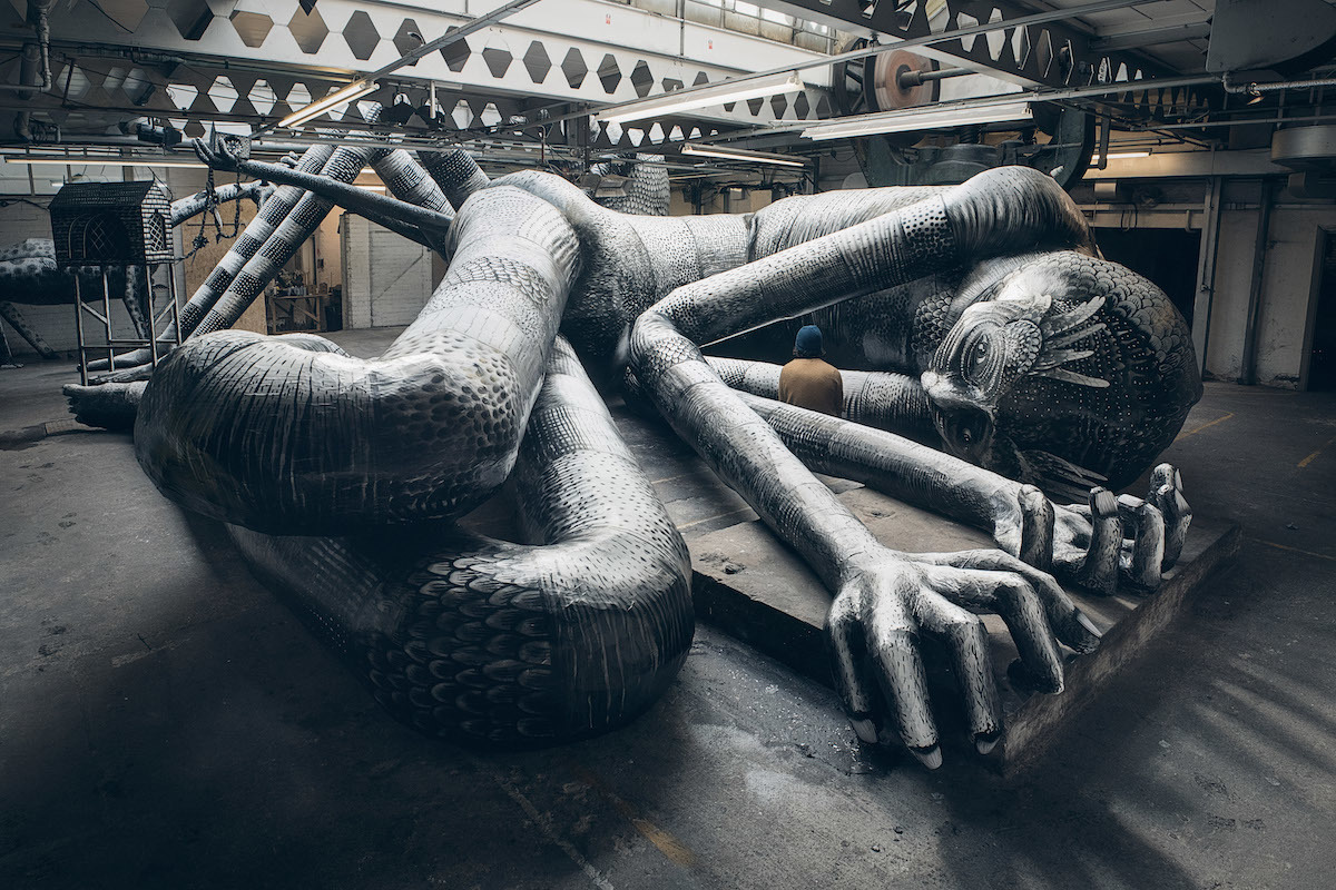 A large, intricate sculpture of a reclining humanoid figure with elongated limbs and detailed patterns covering its surface lies on the floor of an industrial-style warehouse. The artwork features exaggerated hands and feet, a textured body, and a stylized head with decorative markings. Overhead, metal beams and geometric cutouts line the ceiling, while machinery and pipes are visible in the background.