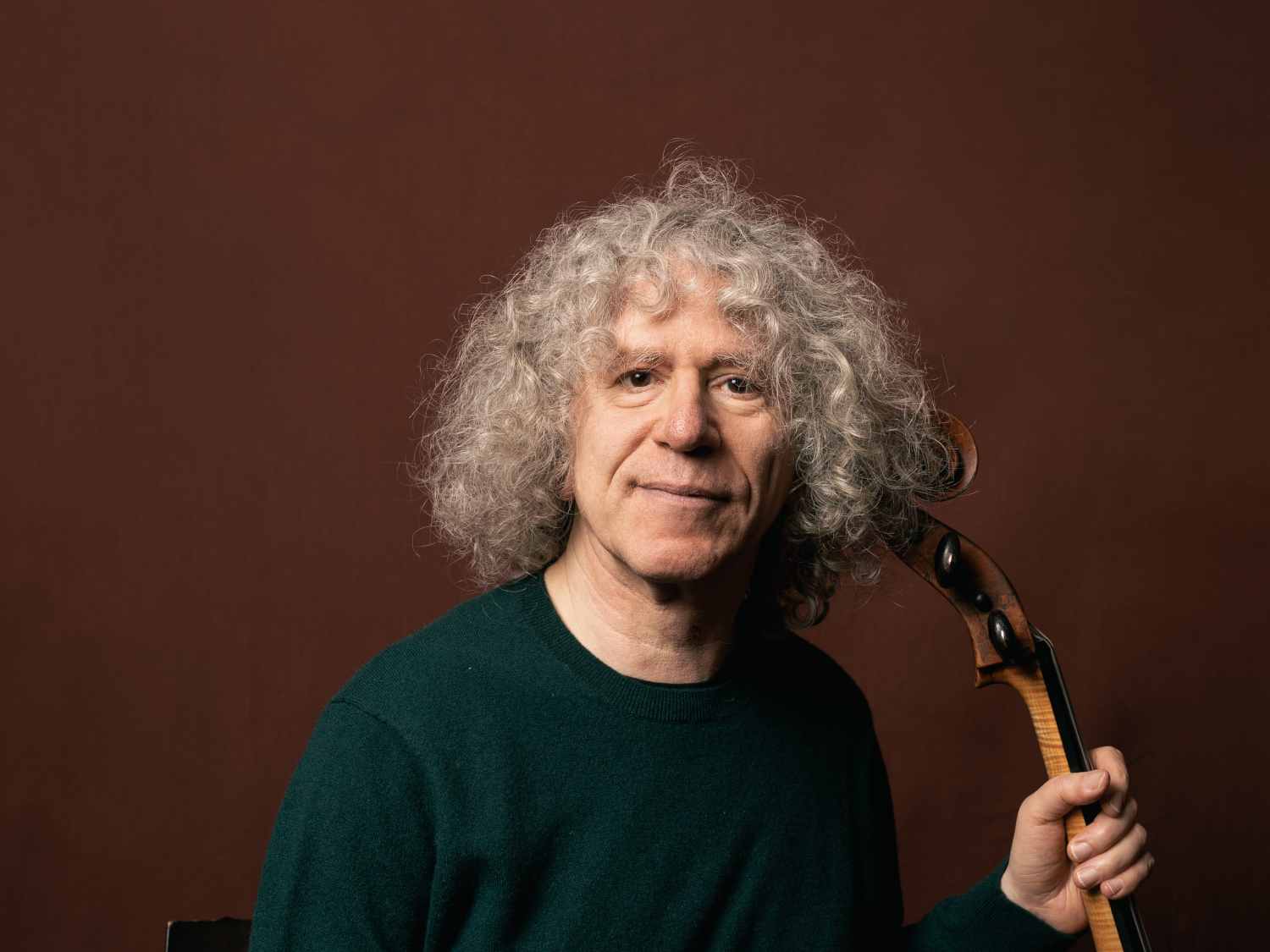 A man, with long, grey curly hair, is sat holding a cello.