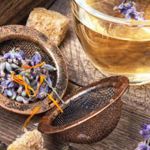 A rustic wooden surface holds a metal tea infuser filled with dried lavender buds and small orange petals, sitting open beside scattered herbs and sugar cubes. Next to it is a clear glass cup filled with light golden herbal tea, with a sprig of lavender resting on the rim. Additional fresh lavender stems lie nearby, creating a warm, natural, and aromatic scene.