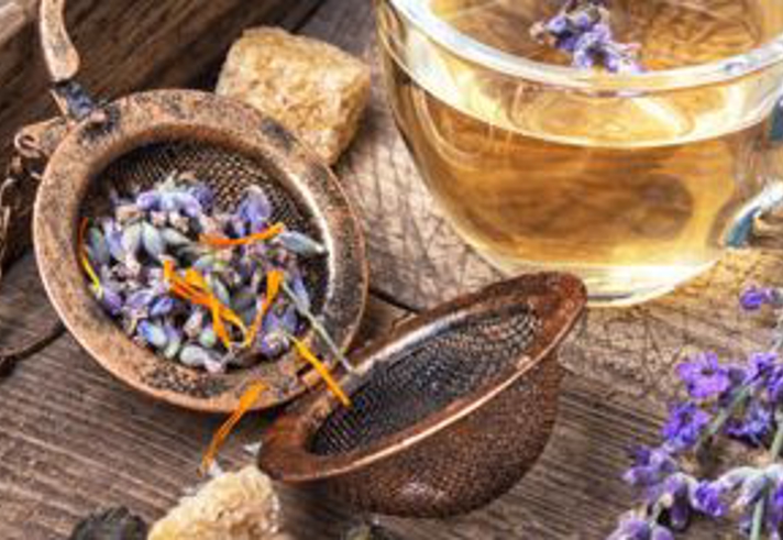 A rustic wooden surface holds a metal tea infuser filled with dried lavender buds and small orange petals, sitting open beside scattered herbs and sugar cubes. Next to it is a clear glass cup filled with light golden herbal tea, with a sprig of lavender resting on the rim. Additional fresh lavender stems lie nearby, creating a warm, natural, and aromatic scene.