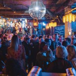 Manahatta filled with people. Crowded bar scene with a large disco ball hanging from the ceiling, surrounded by warm pendant lights and gold balloons. People are gathered closely, chatting and holding drinks. A neon-style sign reading “DON’T WALK” hangs above the crowd, and a chalkboard menu on the right advertises drink and food specials. The atmosphere is lively with dim, colourful lighting and industrial-style decor.