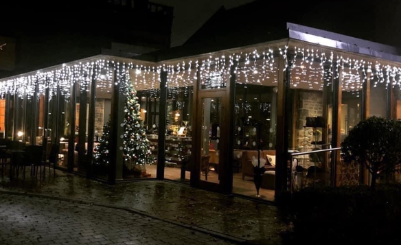 Exterior view of a glass-fronted restaurant at night, decorated with white icicle-style string lights along the roofline. Inside, a Christmas tree with ornaments and lights is visible near the entrance, and warm interior lighting creates a cozy atmosphere. The building is surrounded by a paved walkway and some shrubs, with dark silhouettes of other structures in the background.