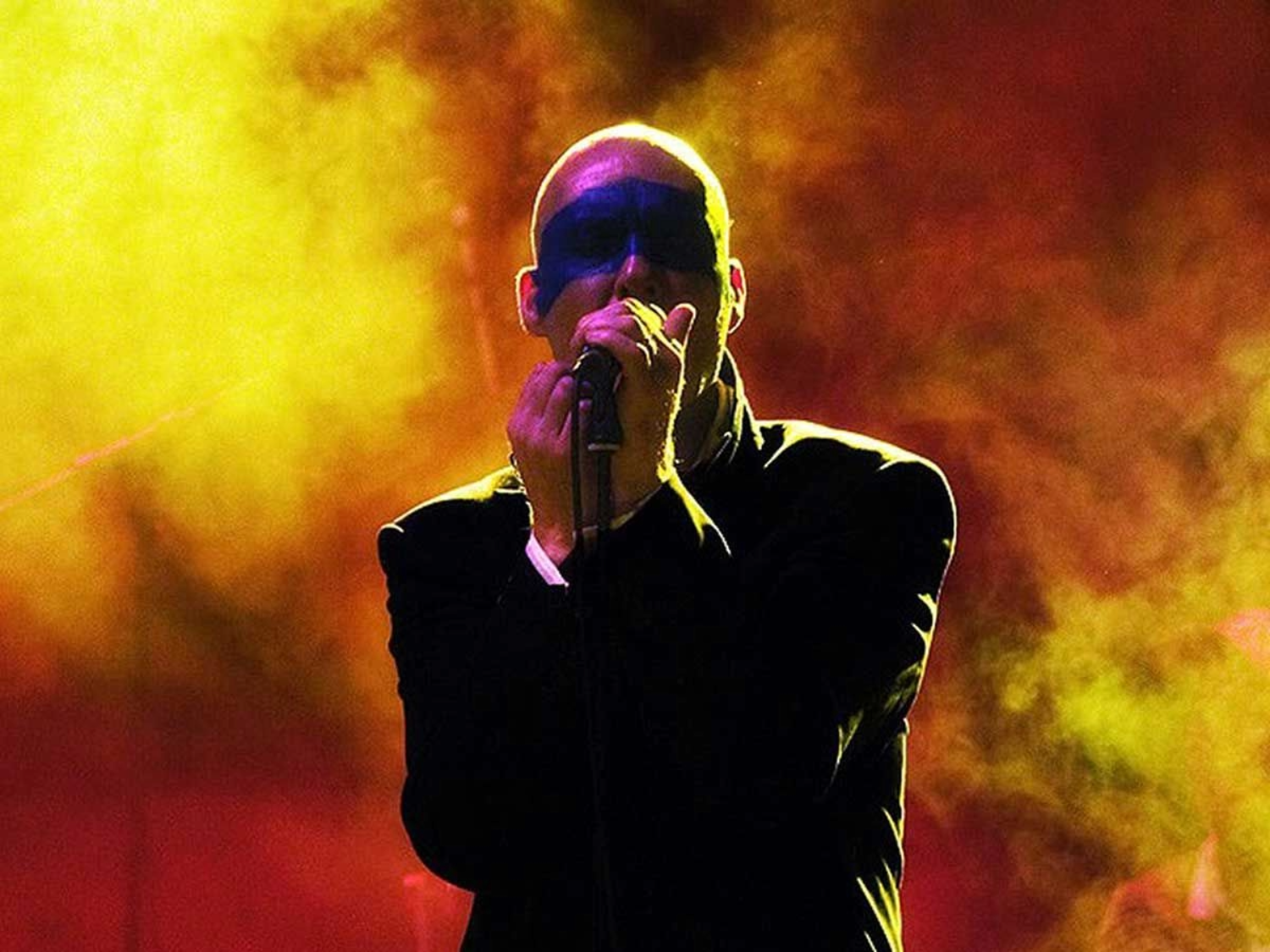 A performer stands on stage holding a microphone close to their mouth, silhouetted against a dramatic backdrop of swirling yellow and red stage lights. The figure is dressed in a dark outfit, creating a striking contrast with the vibrant, smoky background.