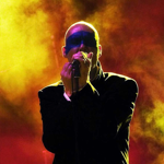 A performer stands on stage holding a microphone close to their mouth, silhouetted against a dramatic backdrop of swirling yellow and red stage lights. The figure is dressed in a dark outfit, creating a striking contrast with the vibrant, smoky background.