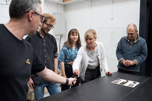 Five people gathered around a black table in a room with white brick walls, engaged in an informal discussion. One person is handing an object to another. Printed materials are on the table, and a whiteboard with text is visible in the background.