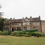 A large historic stone building with multiple windows and a pitched roof, surrounded by lush greenery and landscaped gardens. The structure has a rustic appearance with weathered stone walls and a mix of rectangular and bay windows. In front, there is a well-kept lawn bordered by tall ornamental grasses and shrubs, with mature trees framing the scene under an overcast sky.