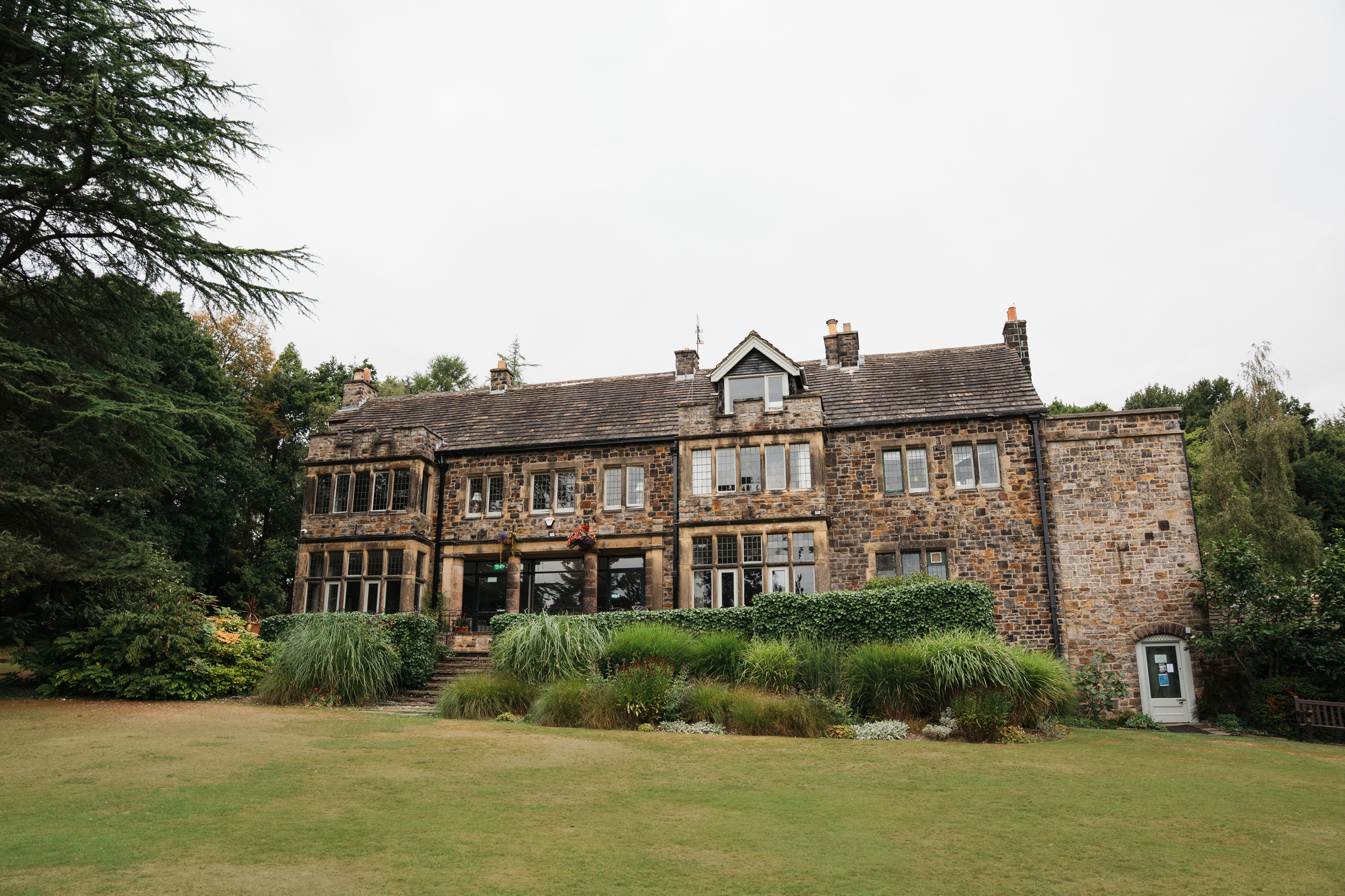 A large historic stone building with multiple windows and a pitched roof, surrounded by lush greenery and landscaped gardens. The structure has a rustic appearance with weathered stone walls and a mix of rectangular and bay windows. In front, there is a well-kept lawn bordered by tall ornamental grasses and shrubs, with mature trees framing the scene under an overcast sky.
