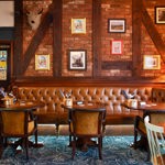 A cosy pub seating area with brick walls, leather banquette seating, round wooden tables, upholstered chairs and framed pictures hung along timber beams.