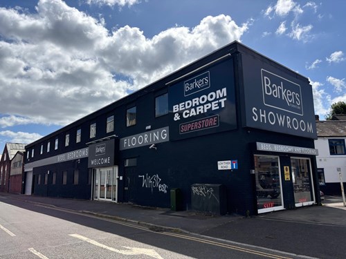 Street view of a large black building with signage reading ‘Barkers Bedroom & Carpet Superstore,’ ‘Barkers Showroom,’ and ‘Flooring.’ The building has multiple windows, a glass entrance door, and a corner display window. Graffiti is visible on the lower wall, and the sky above is partly cloudy with bright sunlight.