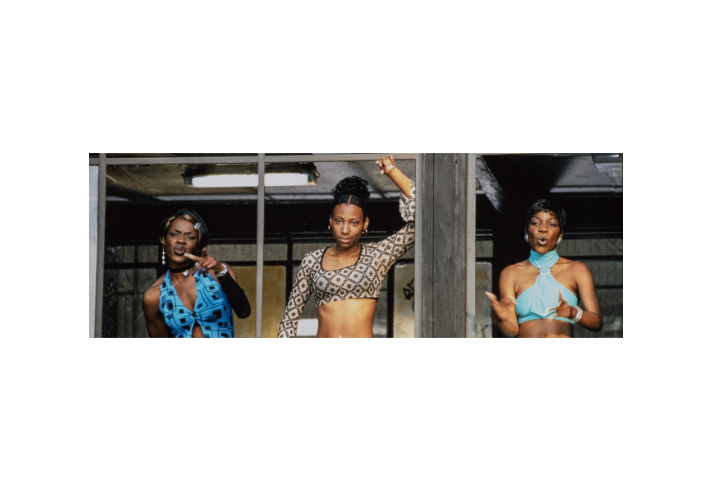 Three people standing in front of a large window frame, dressed in bold, stylish outfits. The person on the left wears a bright blue patterned top with cut-out details and large hoop earrings. The person in the centre wears a cropped long-sleeve top with a black and white geometric pattern, raising one arm above the head. The person on the right wears a turquoise halter-style top with jewellery. The setting appears urban with muted tones and overhead lighting.