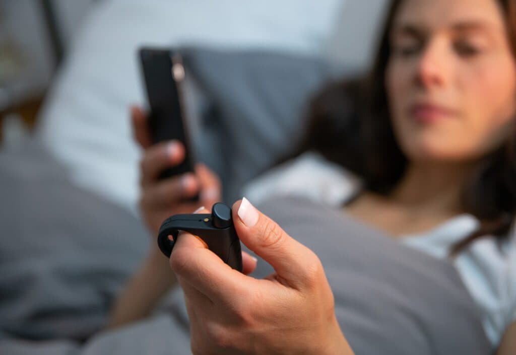 Close-up of a person lying in bed holding a smartphone in one hand and a small black handheld device in the other. The person is covered with a gray blanket, and the background shows pillows and bedding.